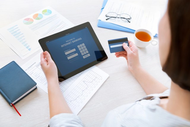 Person using a tablet to access a mobile banking app while holding a debit card, with financial documents and charts on a desk